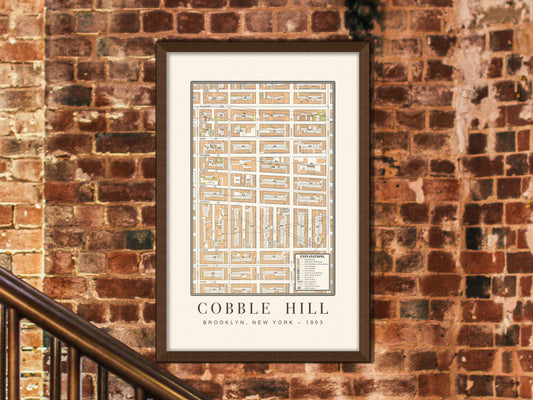 A close-up view of a 1903 historic map of Cobble Hill, Brooklyn, showcased in a dark brown oak frame. The vintage architectural print hangs on a rugged exposed brick wall right beside a metal staircase railing in an industrial-style entryway.