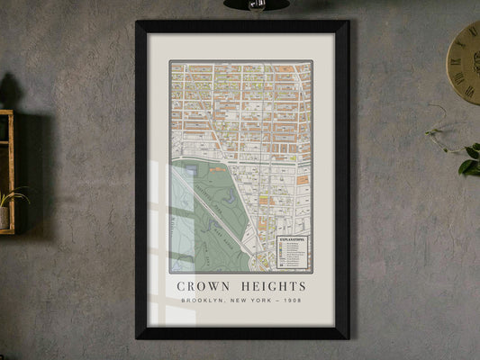 A detailed look at a 1908 vintage map print of Crown Heights, Brooklyn. The archival cartography is displayed in a solid black oak frame on a dark grey textured wall, styled beside a wood shelf featuring a potted trailing vine.