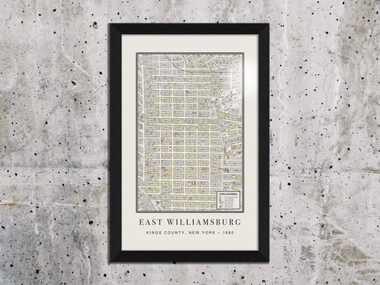An 1880 historical map print of East Williamsburg, Brooklyn, displayed on a textured grey concrete wall. The archival cartography is housed in a solid oak frame with a black finish, complementing a raw industrial interior.