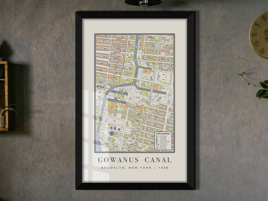 A close-up shot of a 1908 industrial map of the Gowanus Canal in Brooklyn. The premium restored print is framed in solid black oak against a dark grey wall, styled alongside a wooden shelf displaying a hanging green plant.