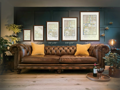 A scale comparison for the vertical 1908 Gowanus Canal map print showcasing five sizes: 12 by 18 inches, 16 by 24 inches, 20 by 30 inches, 24 by 36 inches, and 30 by 45 inches. The industrial Brooklyn cartography is framed in solid dark oak and positioned on a dark paneled wall over a brown leather chesterfield sofa.