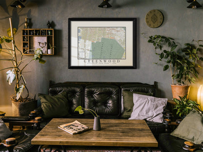 An archival 1908 map of Green-Wood Cemetery in Brooklyn, presented in a solid black oak frame on a dark grey wall. The historic cartography sits above a black tufted leather sofa and a rustic coffee table with surrounding green plants.