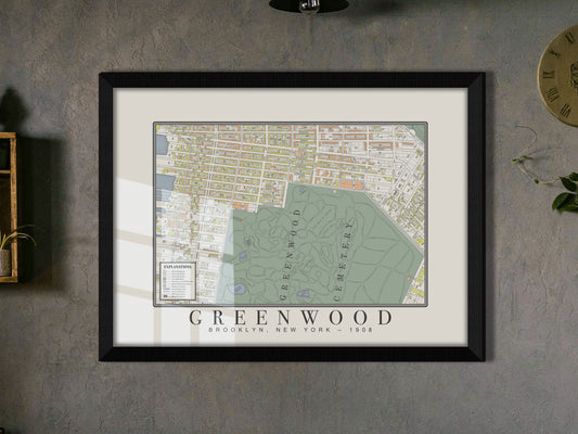 A zoomed-in angle of a 1908 antique map showing Green-Wood Cemetery in Brooklyn. The restored print is mounted in a solid black oak frame against a moody dark grey wall, resting near a rustic wood shelf with a small leafy plant.