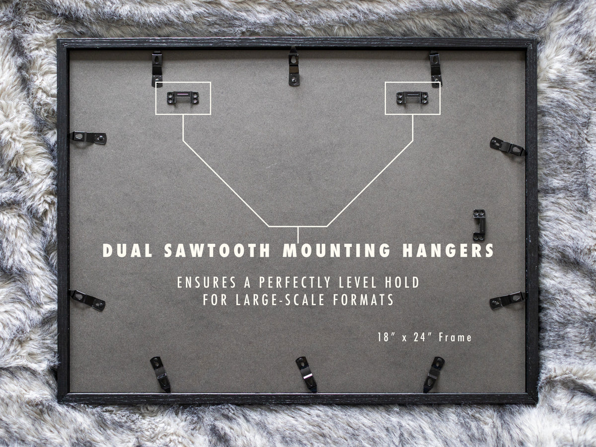 A view of the black backing on a horizontally oriented 18 by 24 inch frame laying on a grey faux fur surface. A white diagram illustrates two metal sawtooth hangers installed at the top corners to support large landscape artwork evenly.