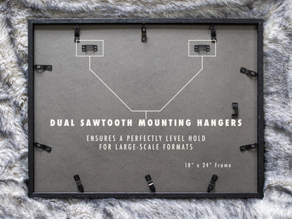 A view of the black backing on a horizontally oriented 18 by 24 inch frame laying on a grey faux fur surface. A white diagram illustrates two metal sawtooth hangers installed at the top corners to support large landscape artwork evenly.