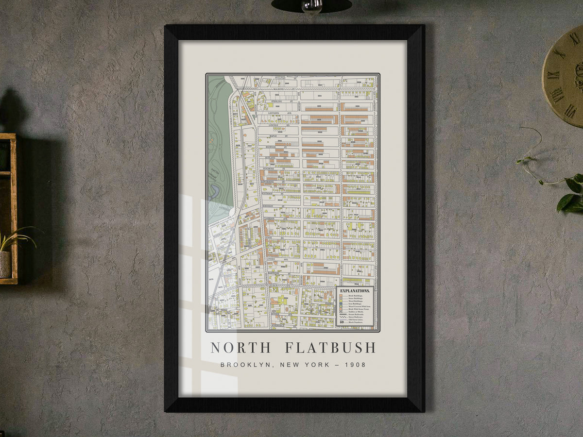 A close perspective of a 1908 historical map of Flatbush, Brooklyn. The framed cartography features a solid black oak frame and hangs on a textured charcoal grey wall, located adjacent to a wooden shelf holding a cascading plant.