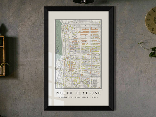 A close perspective of a 1908 historical map of Flatbush, Brooklyn. The framed cartography features a solid black oak frame and hangs on a textured charcoal grey wall, located adjacent to a wooden shelf holding a cascading plant.