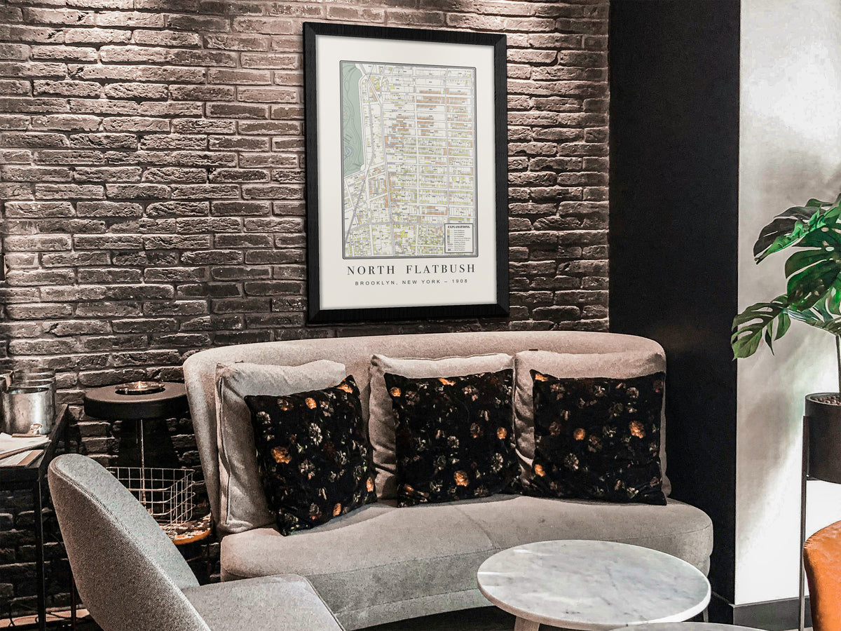 An archival 1908 map print of Flatbush, Brooklyn, mounted on a distressed grey brick wall. The vintage artwork features a solid black oak frame and is positioned over a modern grey couch adorned with dark throw pillows.