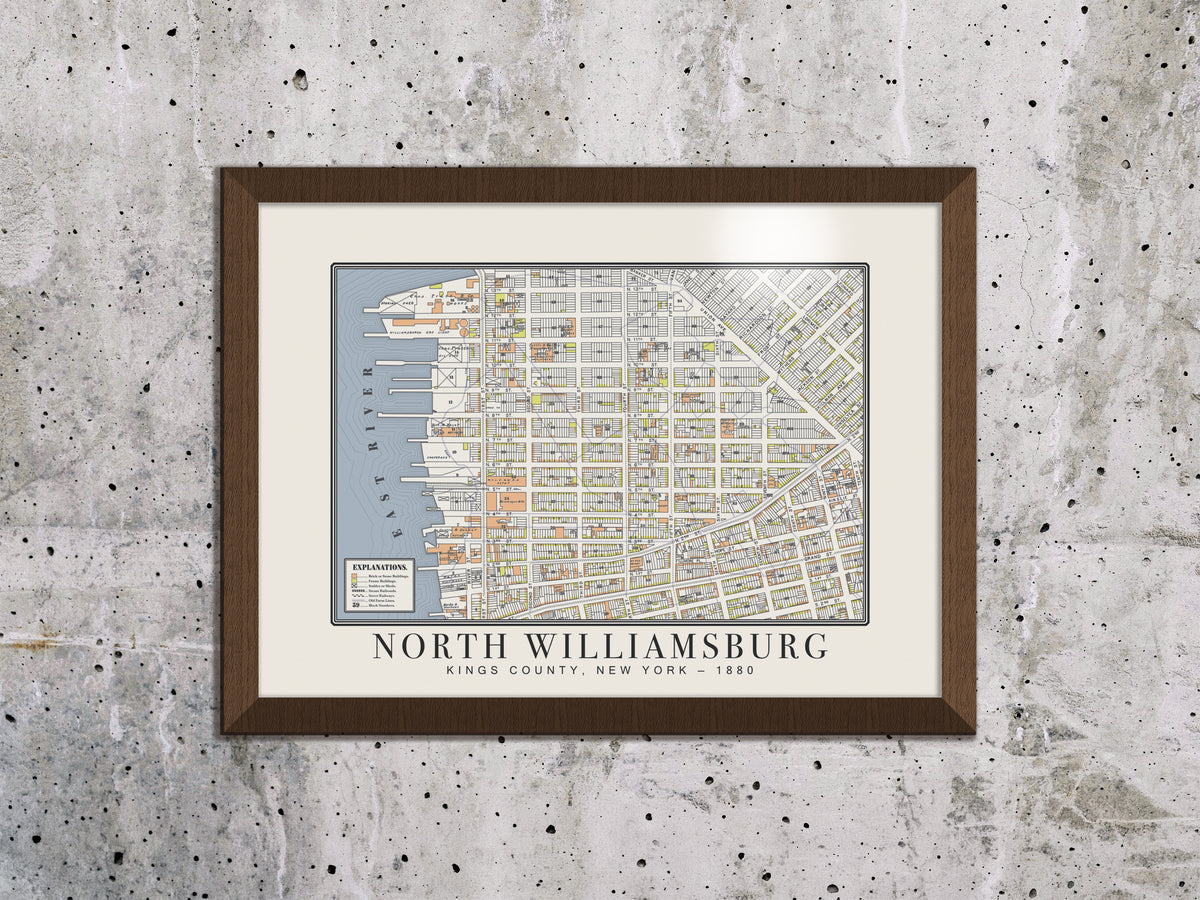 An 1880 antique map of North Williamsburg, Brooklyn, positioned on a raw, distressed concrete wall. The restored survey print is framed in a dark brown oak finish, creating a stark architectural look for modern spaces.