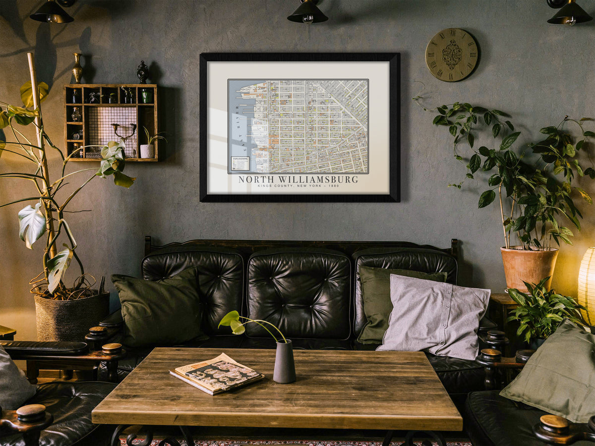 A vintage 1880 map of North Williamsburg, Brooklyn, set in a solid black oak frame against a dark grey textured wall. The map print is centered over a black tufted leather sofa, a wood coffee table, and green house plants.