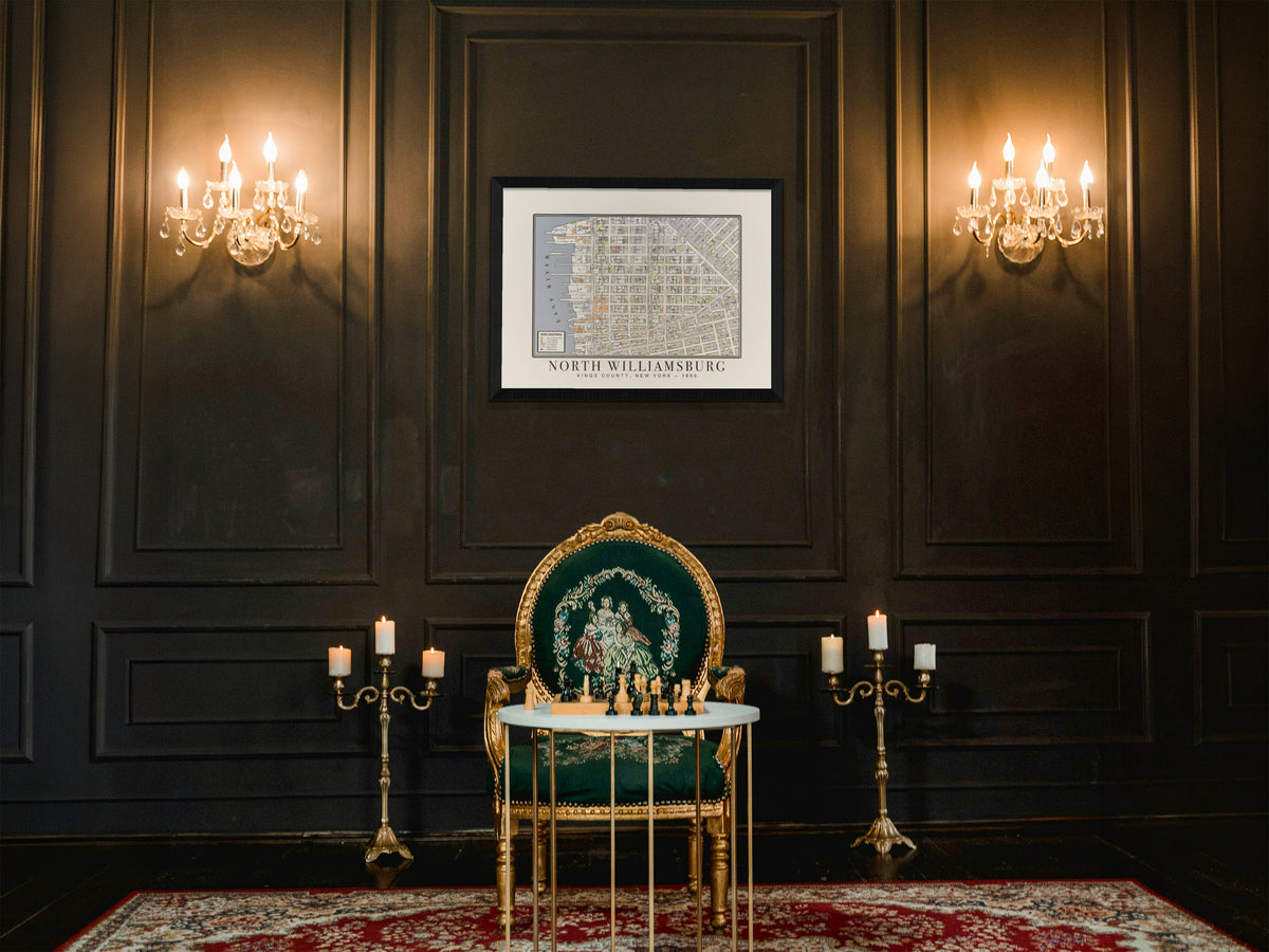 An archival 1880 map of North Williamsburg, Brooklyn, elegantly framed in solid oak on a dark paneled wall. The vintage New York print completes a traditional room setup featuring a green upholstered Victorian chair, a wooden chess board, and glowing candelabras.