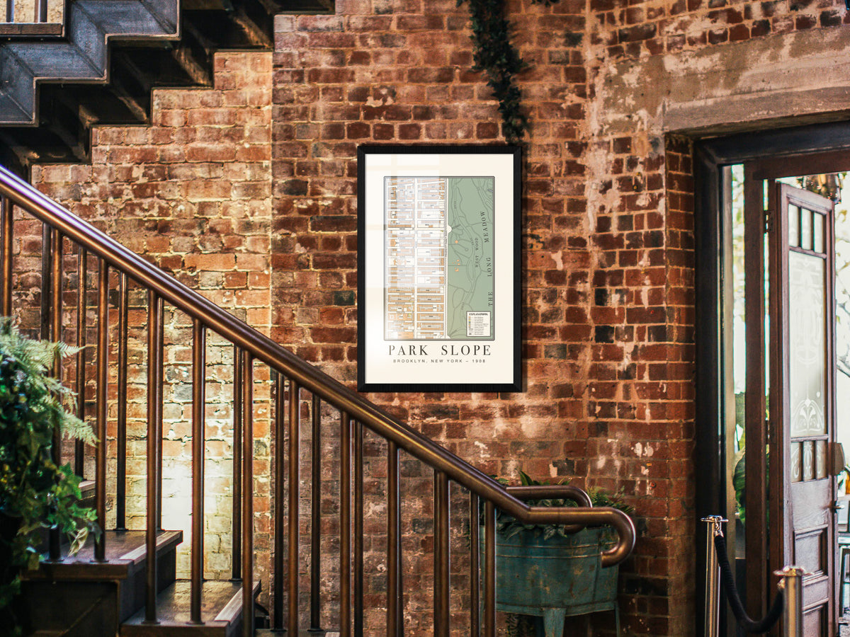 An archival 1908 map of Park Slope, Brooklyn, framed in a solid black oak finish. The historical cartography print hangs on a raw brick wall in a dark industrial entryway space, situated next to a staircase with metal railings.