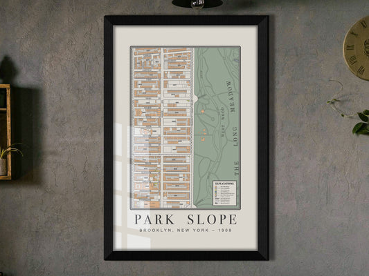 A close-in view of a 1908 vintage map of Park Slope, Brooklyn. The archival print is encased in a solid black oak frame, hung on a textured dark wall, and placed next to a small wooden shelf with a trailing houseplant.