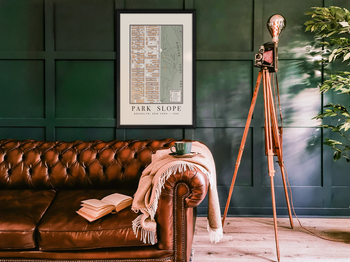 An archival 1908 map print of Park Slope, Brooklyn, encased in a black stained oak frame and mounted on a dark green paneled wall. The historic cartography sits above a brown leather chesterfield couch, a blanket, a coffee mug, and a retro camera floor lamp.
