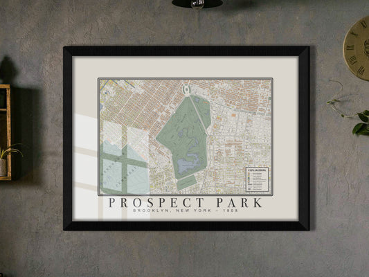A detailed look at an archival 1908 map of Prospect Park, Brooklyn. The historic print is displayed in a solid black oak frame on a moody grey textured wall, situated beside a small wood shelf bearing a trailing green plant.