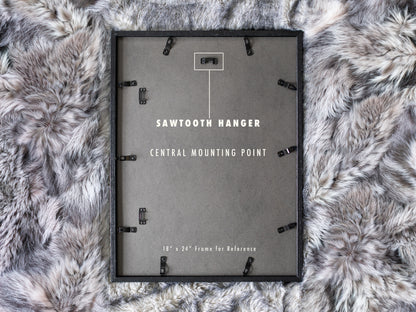 A close-up of the black backing on an 18 by 24 inch portrait-oriented frame lying face down on a faux fur rug. A white diagram points to a single metal sawtooth hanger attached at the top center for level wall mounting.