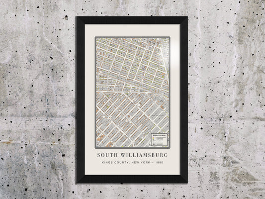 A restored 1880 vintage map of South Williamsburg, Brooklyn, mounted on an unfinished concrete wall. The architectural print features a black oak frame, highlighting an exposed, industrial interior design style.